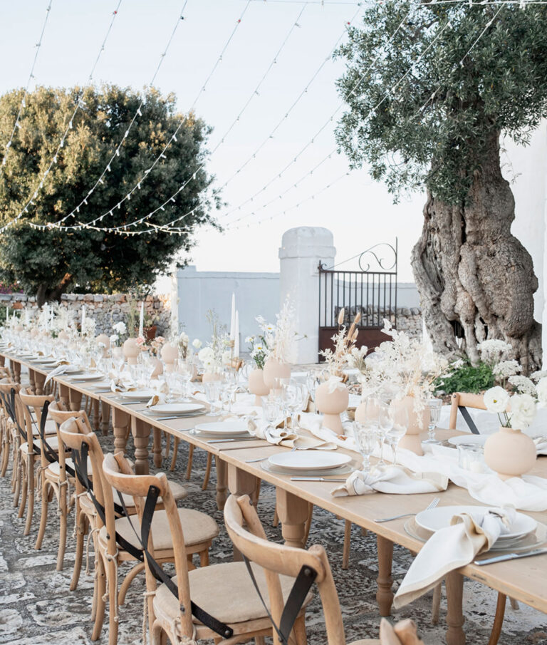 A long table under an old olive tree in a whitewashed masseria in Puglia, neutral flowers and strings of lights overhead, Glam Events in Tuscany