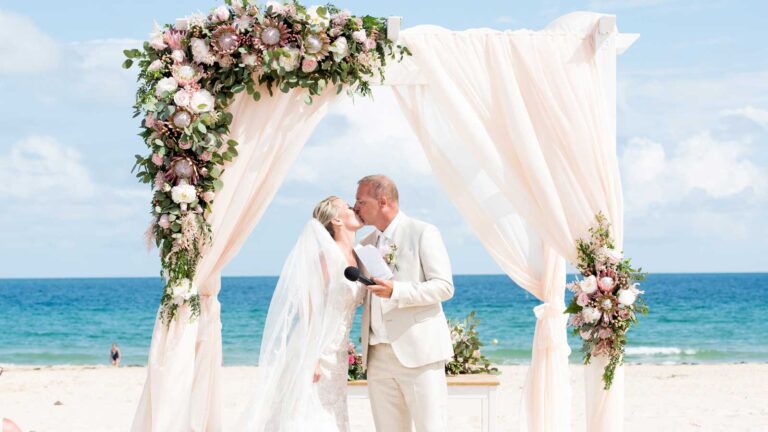 Couple kissing beneath a draped floral arch on the beach, Glamour Algarve Weddings
