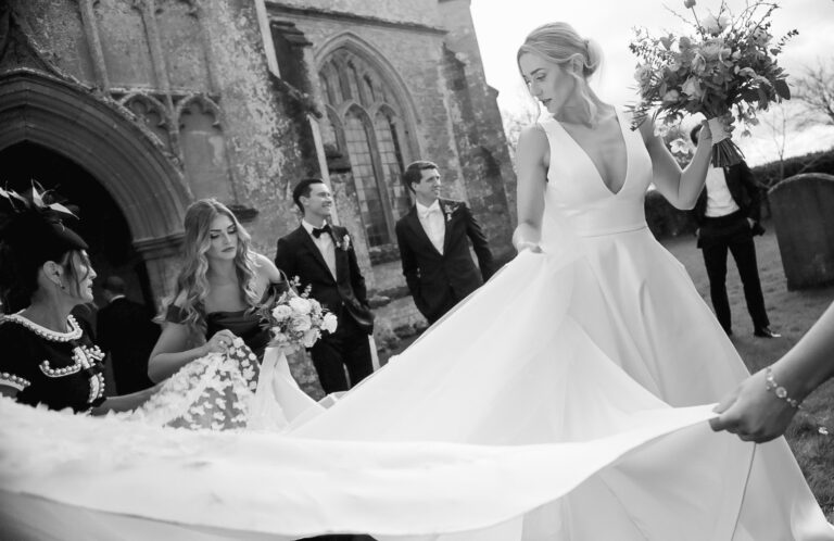 Black and white frame of bridesmaids gathering the bride's train outside a church, Gorgeous Films