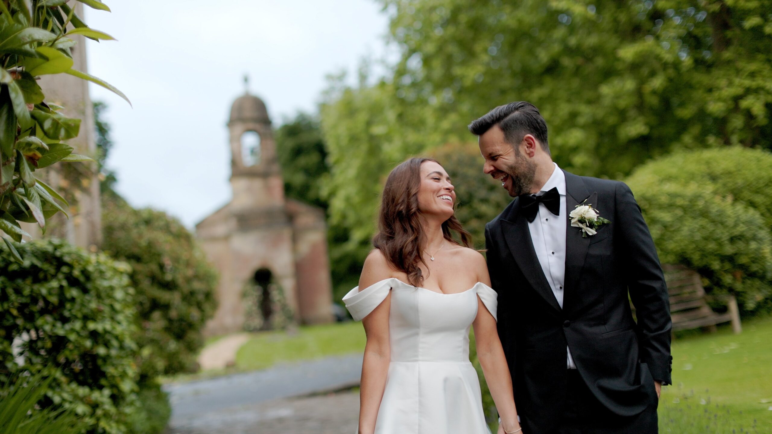 A bride and groom walking through a garden, a small chapel with a bell tower behind them, Gorgeous Films