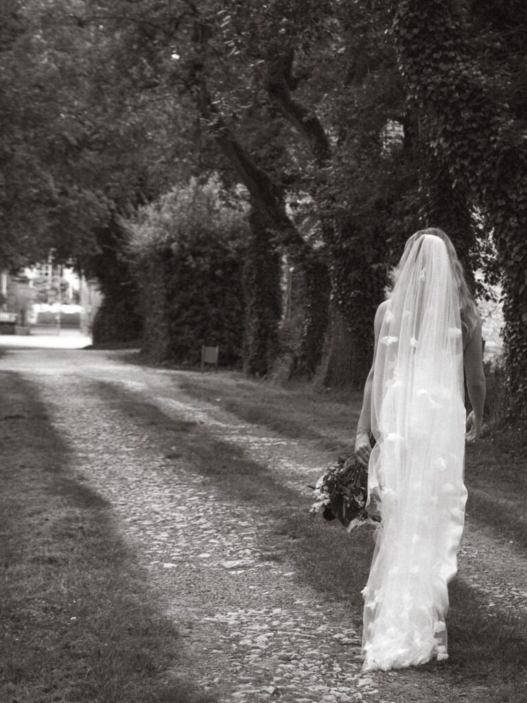 Black and white frame of a bride walking alone down a tree-lined avenue, Gorgeous Films