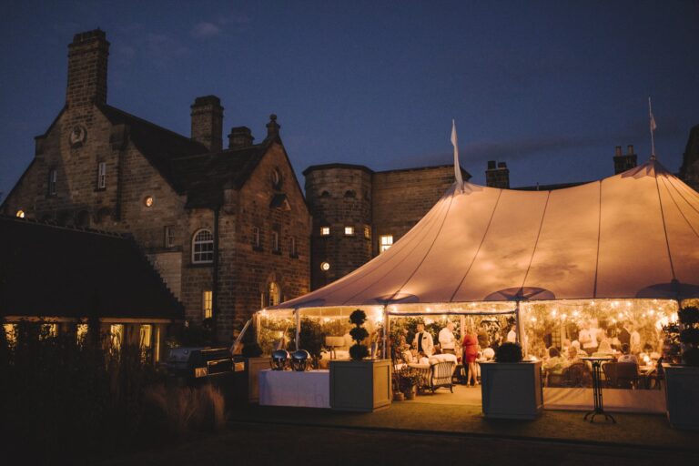 A sailcloth marquee lit up at night beside the older stone wings of the Hall, Grantley Hall