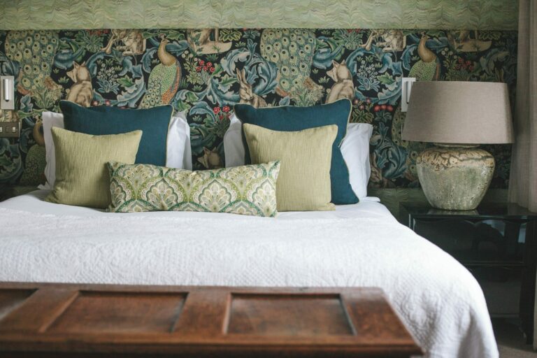 A Manor House bedroom dressed in peacock-patterned Morris wallpaper with teal velvet cushions, Hampton Manor