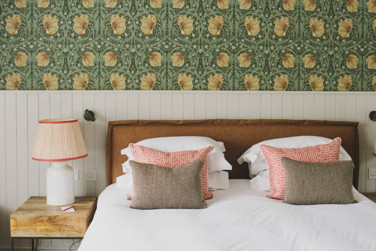 A bedroom in green Morris wallpaper, the bed layered with coral and linen cushions, Hampton Manor