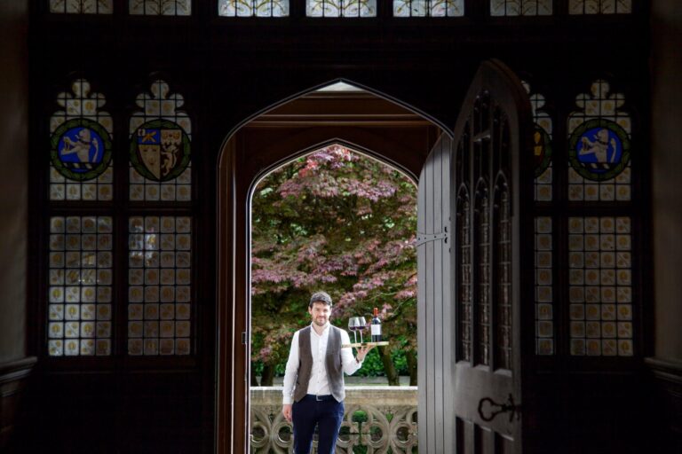 A stained-glass doorway opening to spring blossom, a host waiting in waistcoat, Hampton Manor