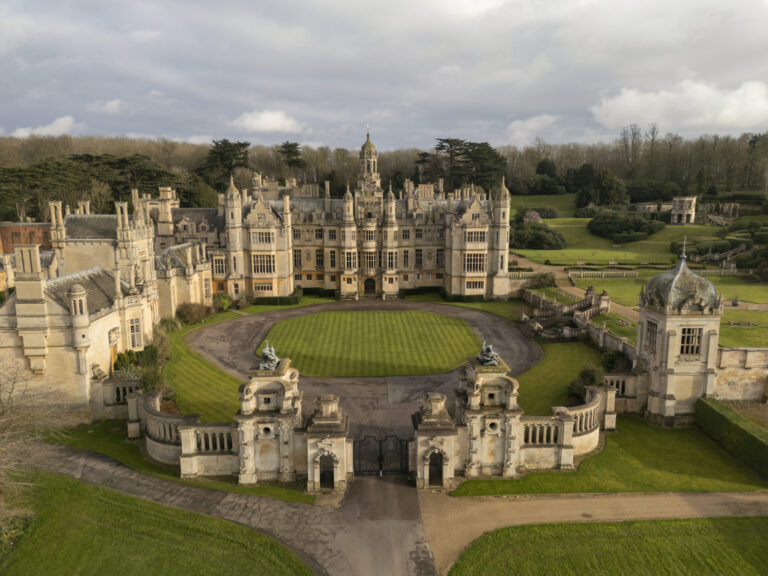The manor, its walled forecourt and gate piers from the air under a grey sky, with parkland and woodland behind, Harlaxton Manor