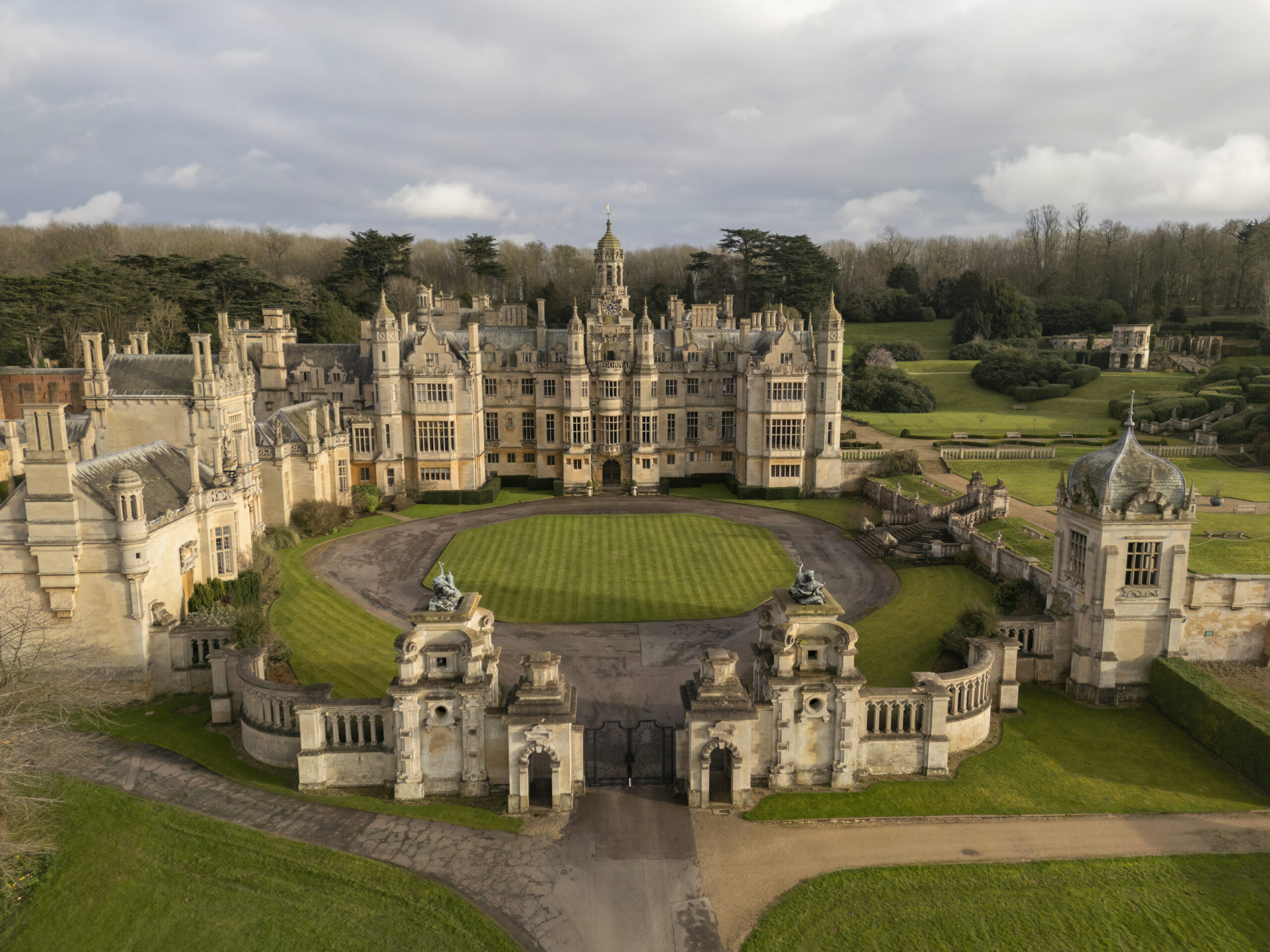 The manor, its walled forecourt and gate piers from the air under a grey sky, with parkland and woodland behind, Harlaxton Manor