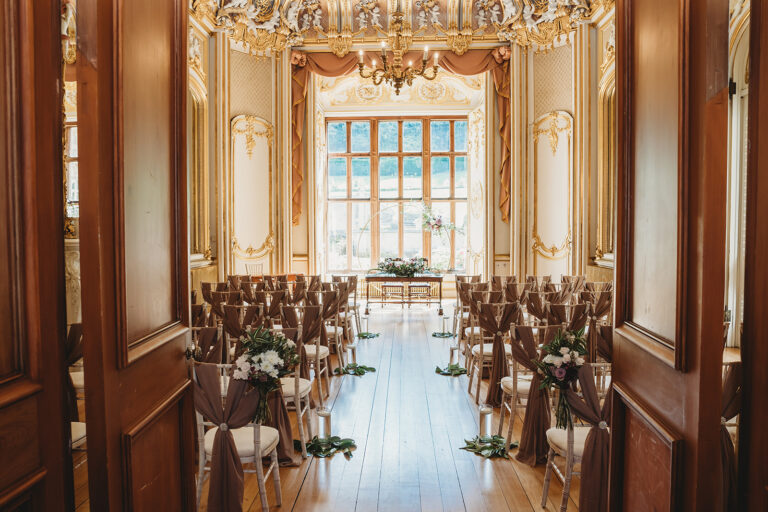 The Gold Room seen through open doors, set for a ceremony with draped chairs, a centre aisle and a table under the bay window, Harlaxton Manor