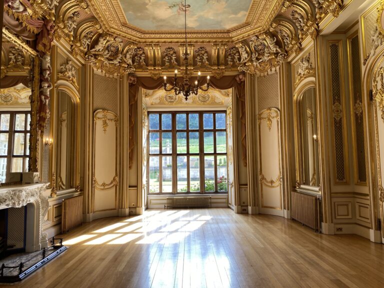 The empty Gold Room in sunlight, with gilded plasterwork, a marble fireplace and a large bay window onto the gardens, Harlaxton Manor