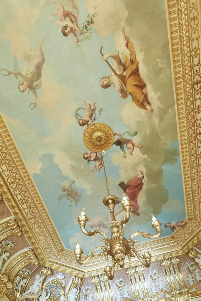 A painted ceiling of cherubs and figures in a sky, framed by gilded plasterwork, with a gilt chandelier, Harlaxton Manor