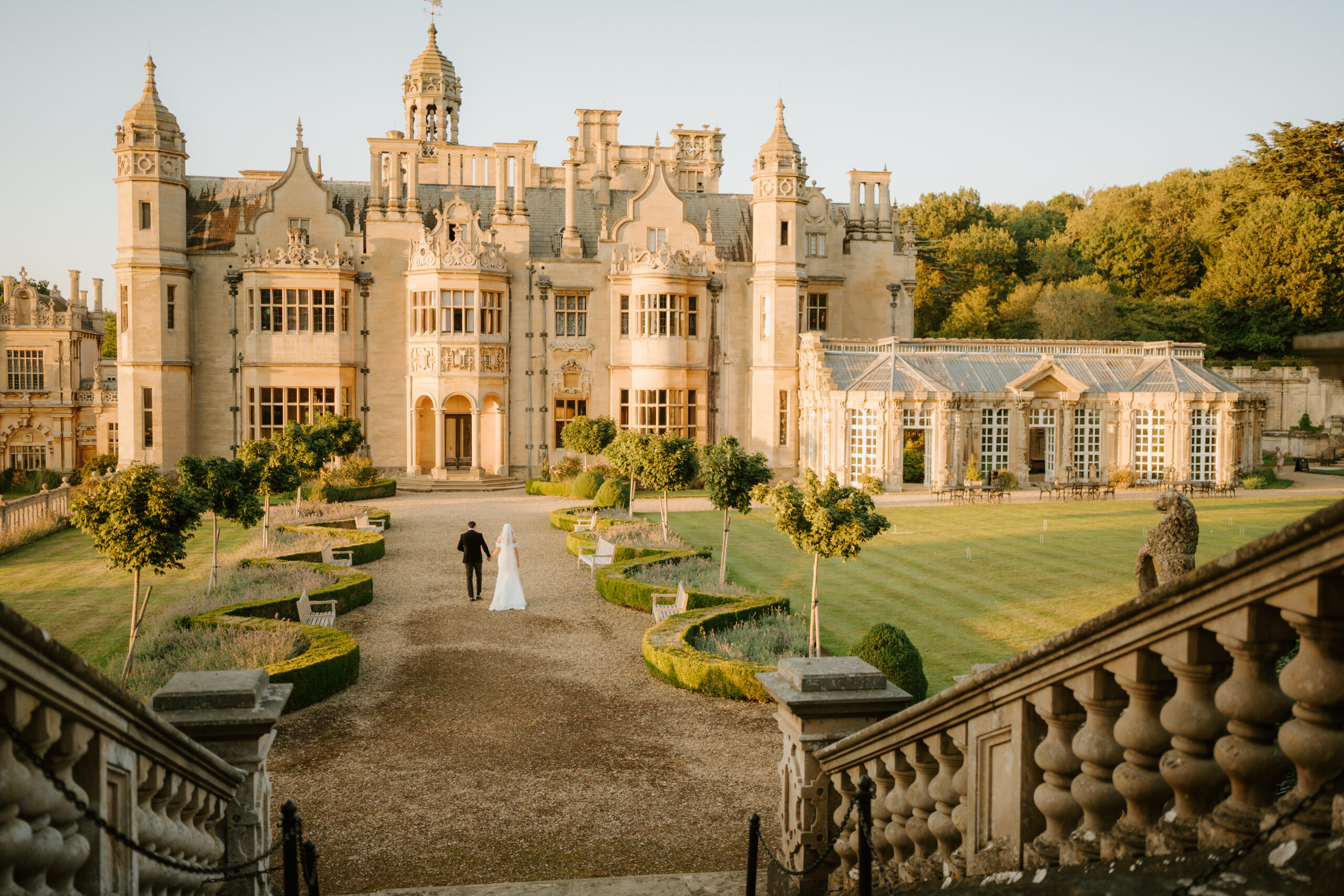 The garden front in evening light, seen from the top of the terrace steps, with a couple walking up the gravel path between clipped hedges, Harlaxton Manor