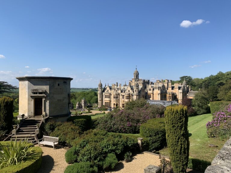 The manor seen from the upper gardens beside a stone garden pavilion, with steps, clipped yew and lawns below, Harlaxton Manor