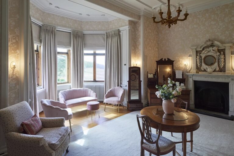 A sitting room inside the manor with a bay window, an open fireplace and pale upholstered chairs, Harlaxton Manor