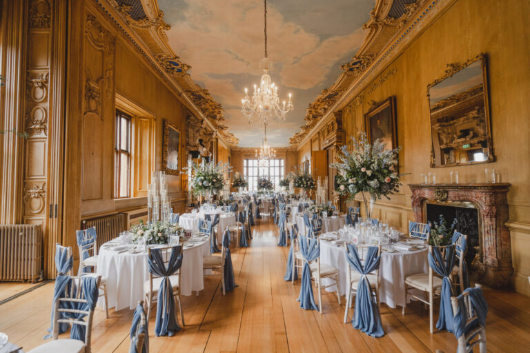 The Long Gallery laid for a wedding breakfast with round tables, blue chair sashes and tall floral arrangements under a painted sky ceiling, Harlaxton Manor