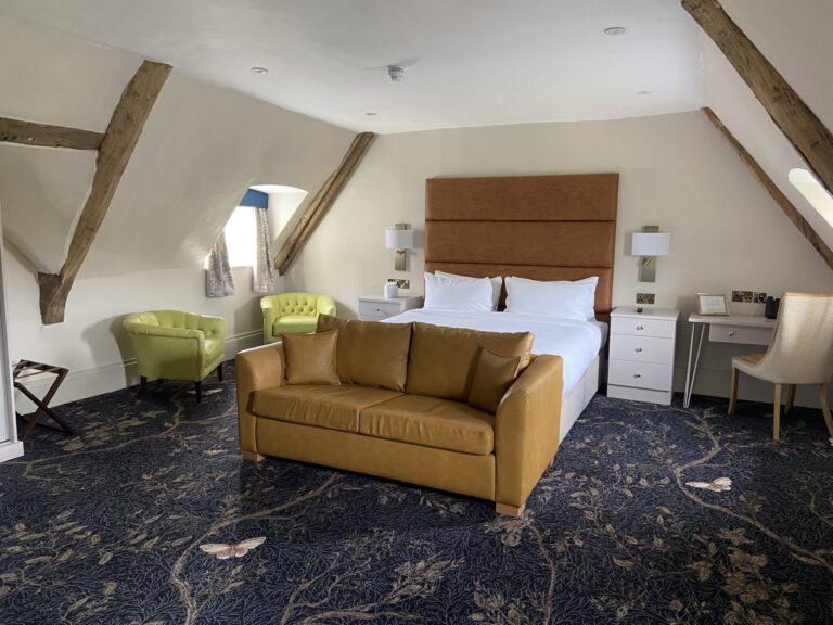 A spacious room under exposed roof timbers with a sofa, armchairs and desk, Haycock Manor