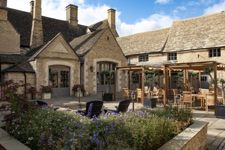 The courtyard terrace behind the hotel, with planted stone beds, pergolas and outdoor tables against the old stone ranges, Haycock Manor