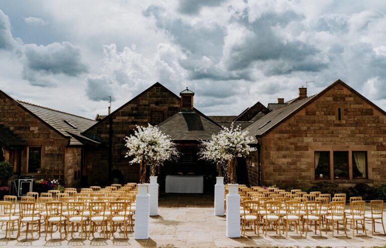 Ceremony set on the patio in front of the oak-framed pagoda, with white blossom trees and gold chairs, Heaton House Farm
