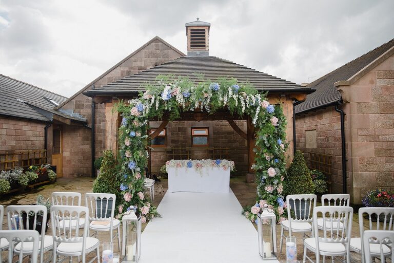 The oak pagoda dressed with a pink and blue floral arch over an ivory aisle and white chairs, Heaton House Farm