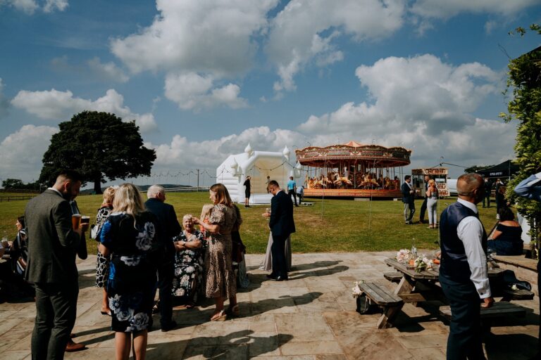 Guests on the patio with a vintage carousel and bouncy castle on the lawn behind, Heaton House Farm