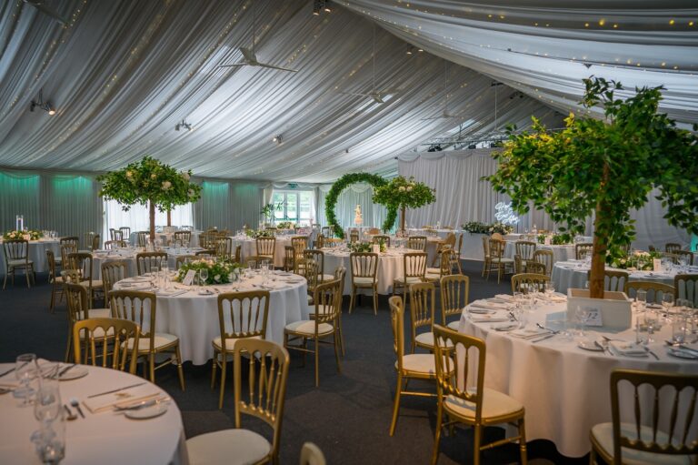 Steeles Barn laid for a wedding breakfast, with white marquee linings and round tables under green-leaved trees, Heaton House Farm