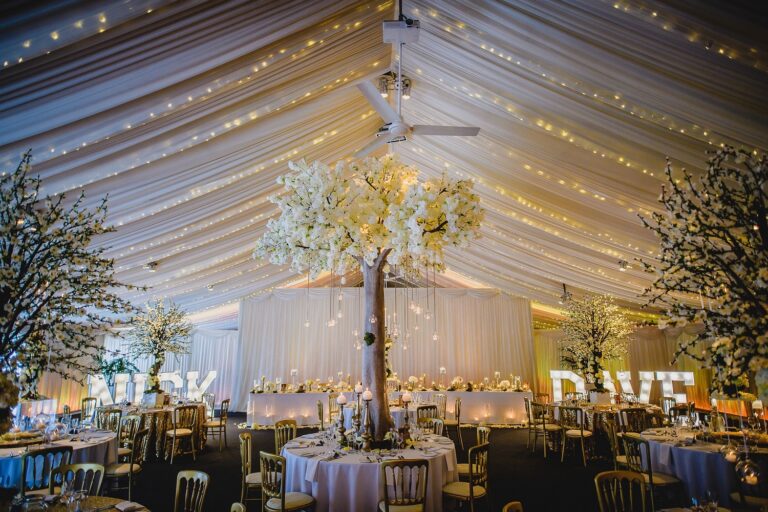 Round tables beneath blossom trees and a fairy-lit drape backdrop in the dining barn, Heaton House Farm