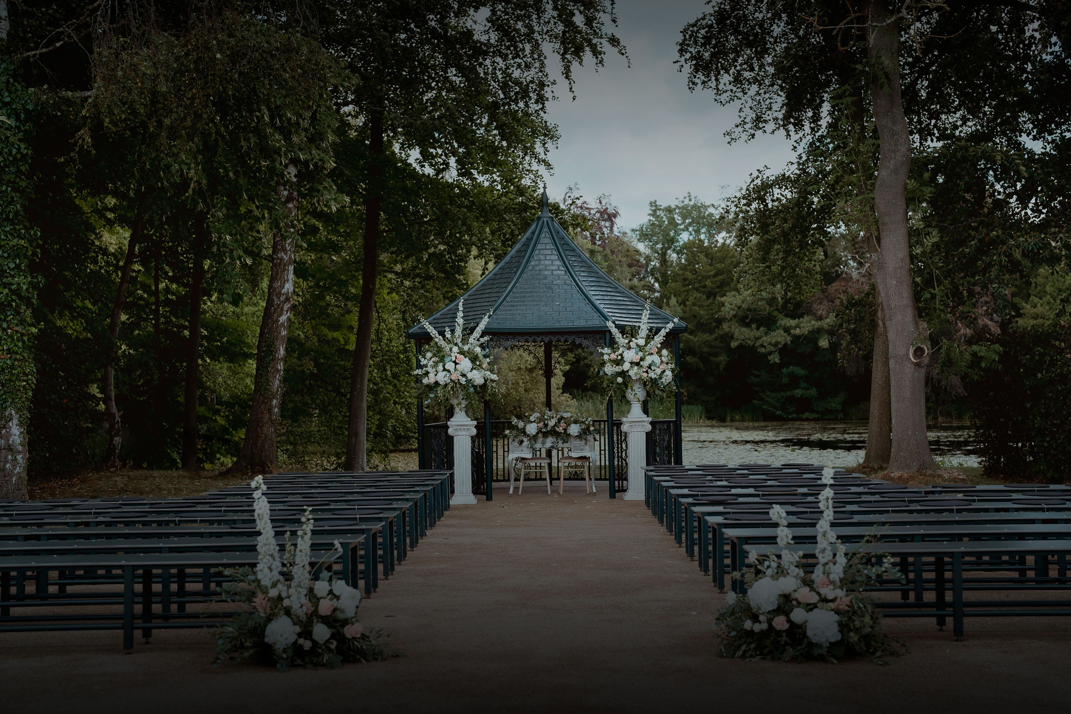 An outdoor ceremony set beneath the trees, an aisle of benches leading to a floral-dressed pavilion, Hengrave Hall