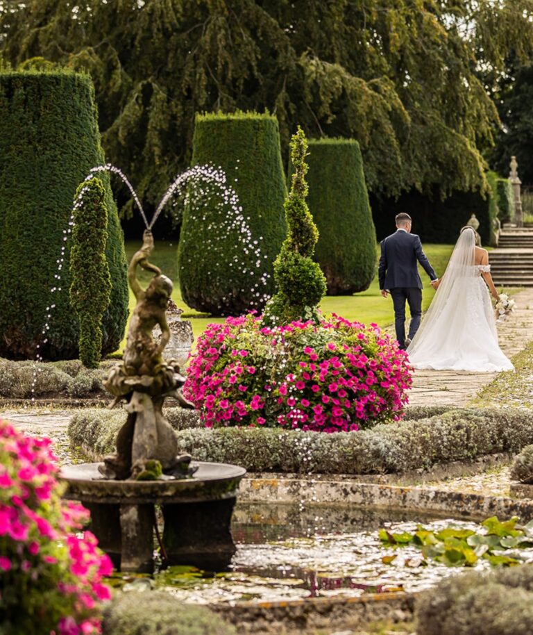 A couple walk through the topiary garden past a fountain and beds of pink flowers, Hengrave Hall