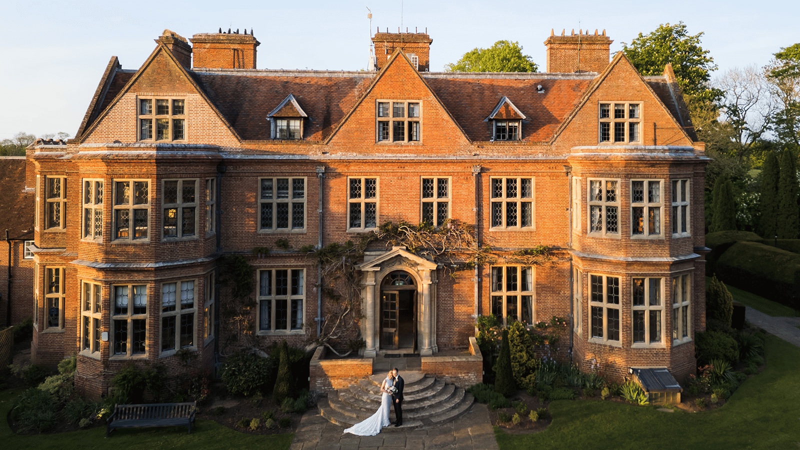 The red-brick Manor House front at golden hour, with a couple on the curved stone steps below the pedimented doorway, Horwood House Hotel & Spa