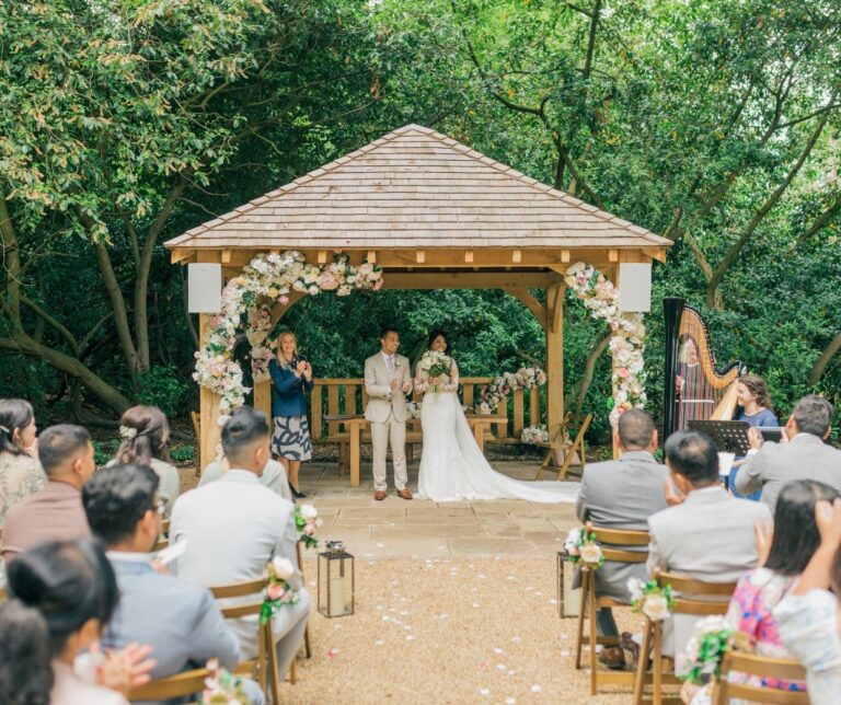 An outdoor ceremony beneath the oak structure, guests seated in the glade, Hylands Estate