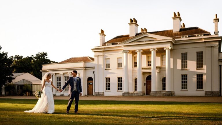 The couple hand in hand on the back lawn at golden hour, the white south front behind them, Hylands Estate