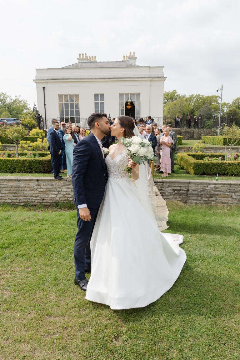 The couple embracing on the lawn, guests and the white facade behind, Hylands Estate