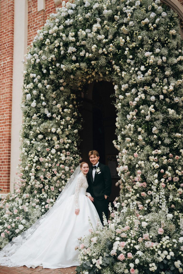 A bride and groom standing in a doorway framed by a tall arch of white and pale pink flowers on a brick facade, IAMFLOWER