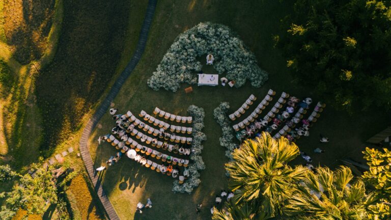 An aerial view of a lawn ceremony with a floral arch and curved rows of chairs among trees, IAMFLOWER