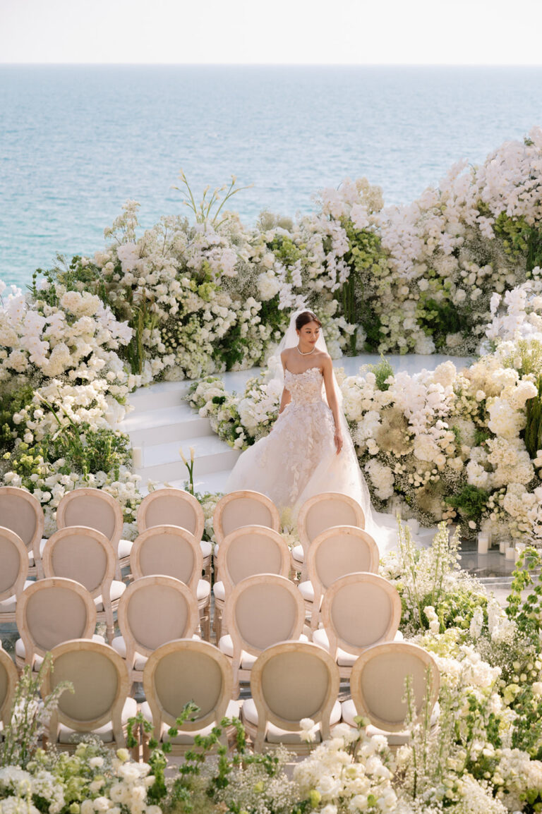 A couple at an altar of white flowers on a lawn above the sea, with rows of guests seated facing them, IAMFLOWER