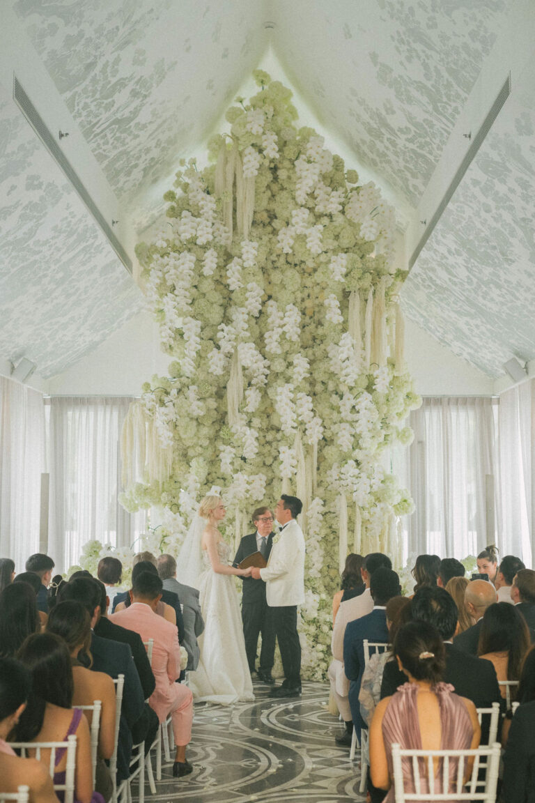 A couple exchanging vows beneath a tall hanging wall of white flowers in a pitched-roof pavilion, with guests seated below, IAMFLOWER