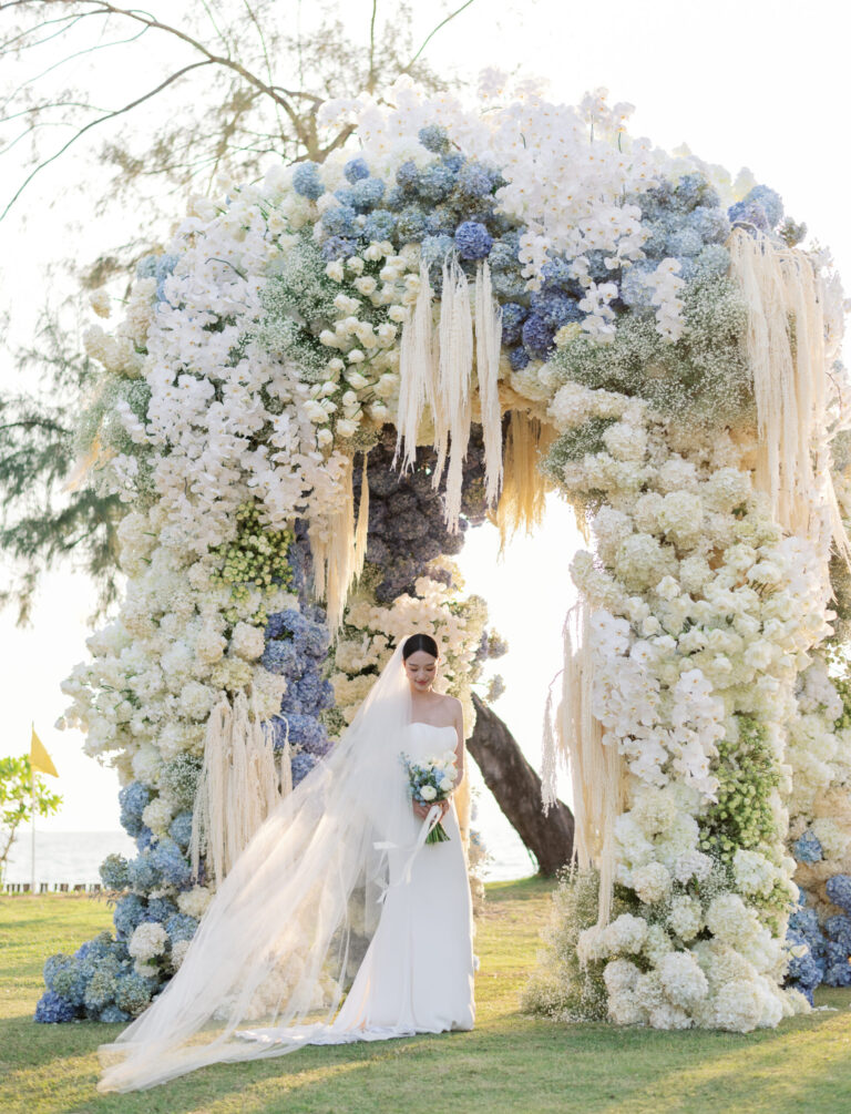 A bride beneath a tall arch of white orchids, blue hydrangeas and trailing amaranthus beside the sea, IAMFLOWER