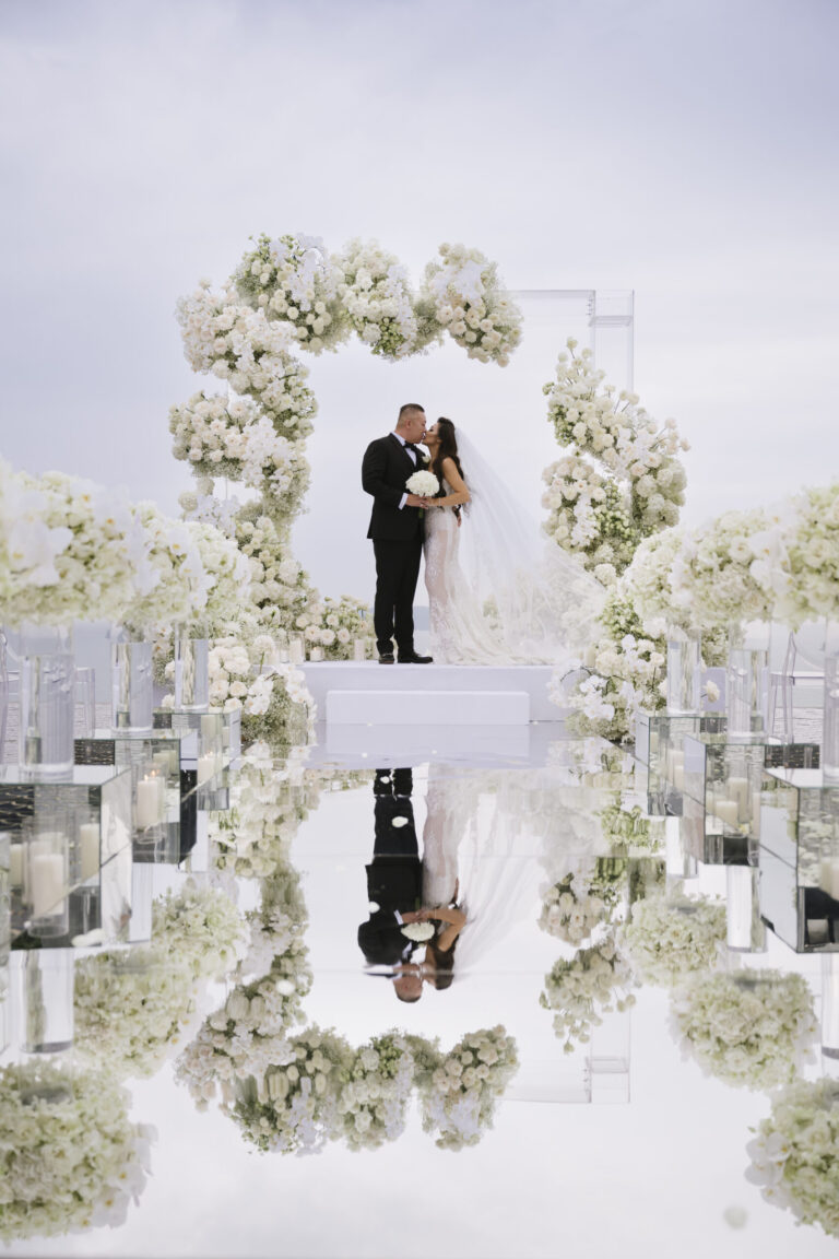 A couple kissing on a white platform framed by a ring of white flowers, with a mirrored aisle and glass candle holders, IAMFLOWER