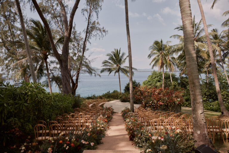 A garden ceremony with wooden chairs and rose-filled borders along a path overlooking the sea through palms, IAMFLOWER