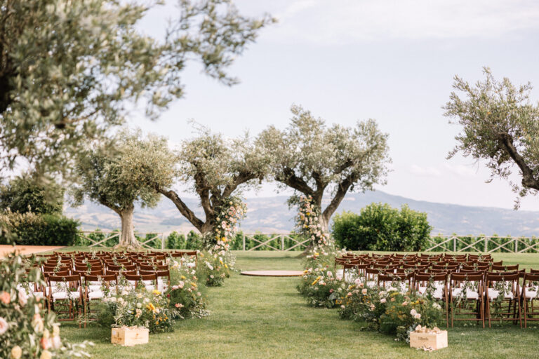 Rows of wooden chairs and flower-lined aisles in an olive grove with hills beyond, a Tuscan ceremony, IAMFLOWER