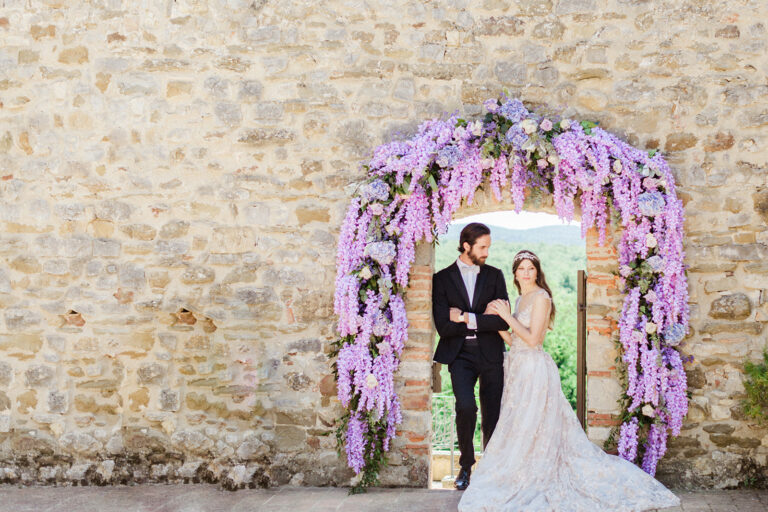 A couple standing in a stone archway wrapped in lilac orchids and wisteria-style blooms, IAMFLOWER