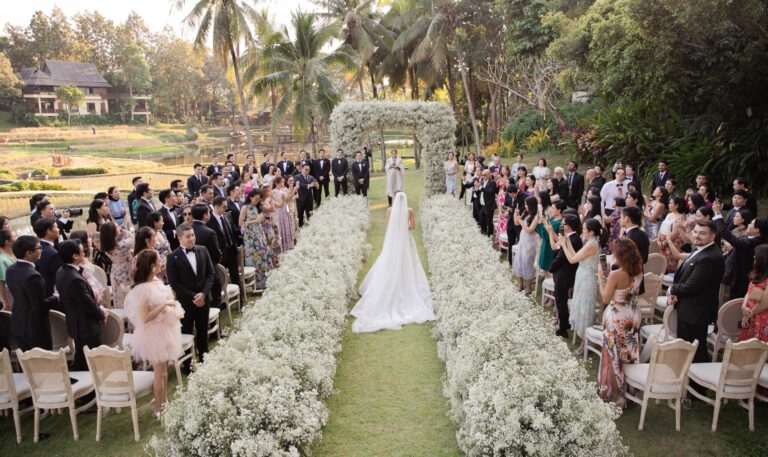 A bride walking a lawn aisle edged with baby's breath towards a floral arch, with guests on both sides, IAMFLOWER