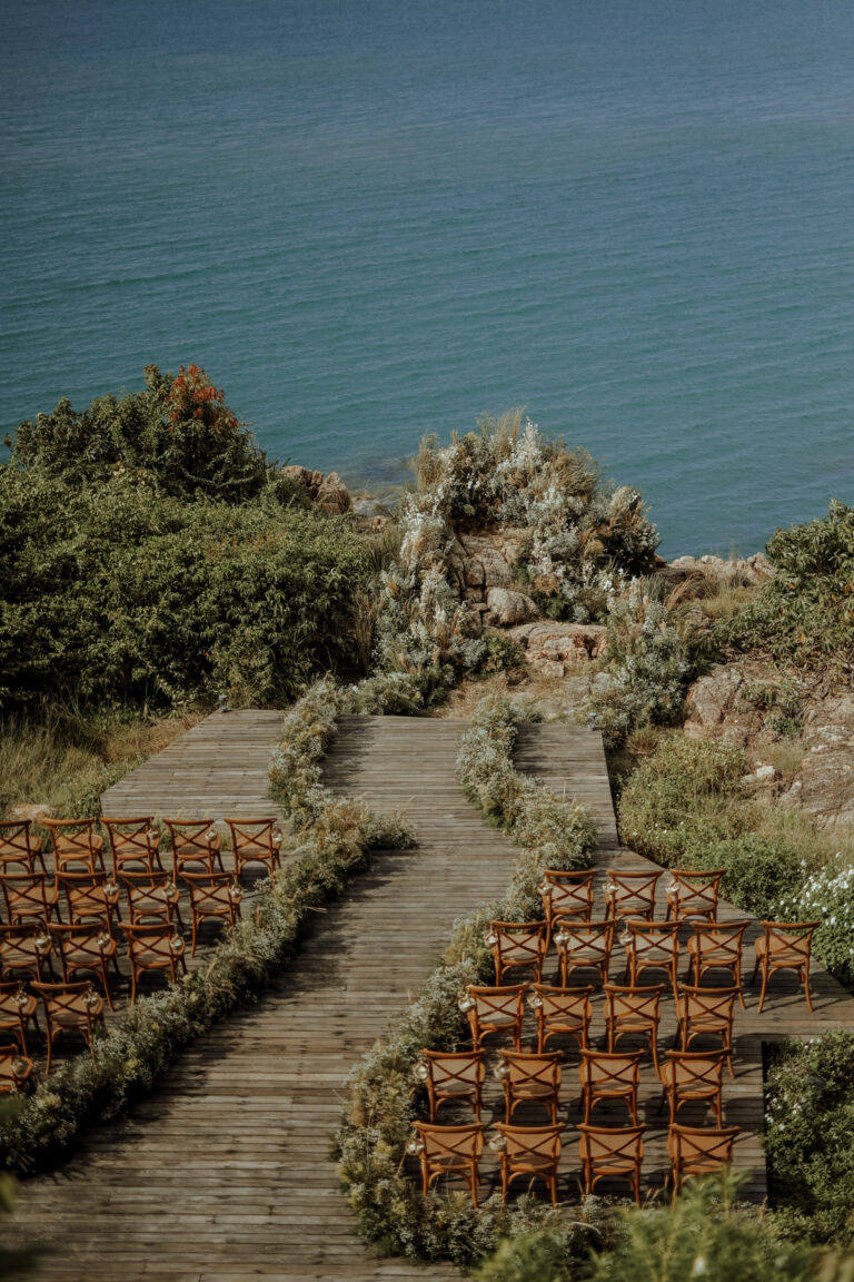 A wooden boardwalk aisle through coastal scrub with rows of wooden chairs, above the sea, IAMFLOWER