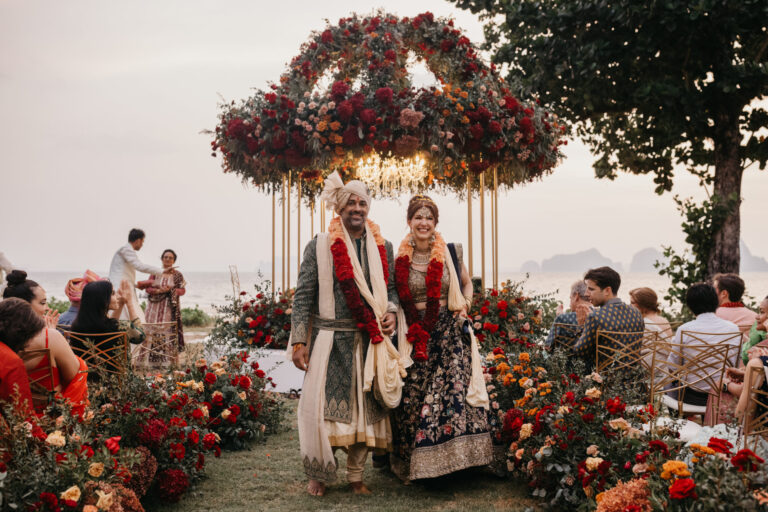 A couple under a gold canopy of red and burgundy flowers, with red blooms around the aisle and the bay behind, IAMFLOWER