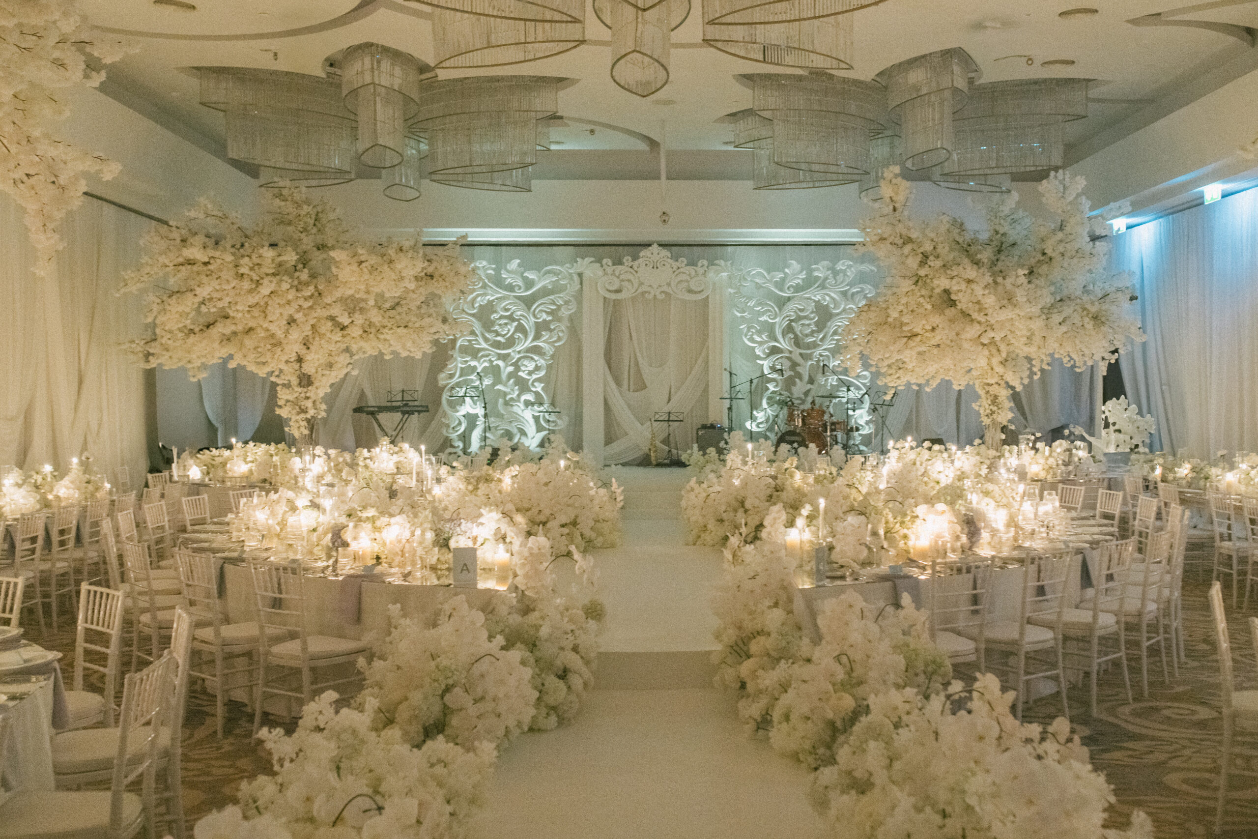 A ballroom reception with round tables, white floral centrepieces and tall floral arrangements in front of a lit backdrop, IAMFLOWER
