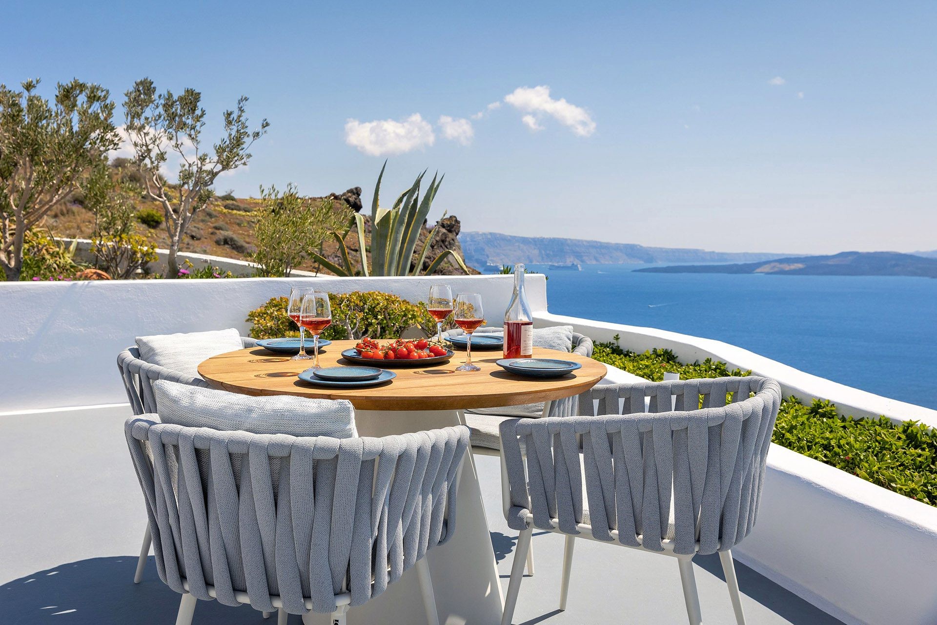 A round table laid with glasses and plates on a terrace, in full sun above the sea