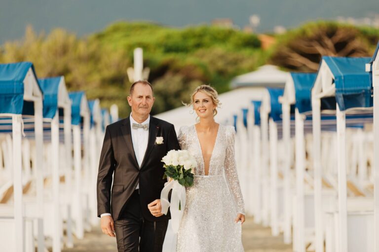 The bride arriving on her father's arm along the boardwalk between blue and white beach cabins, Il Giorno Perfetto di Glenda Marradi