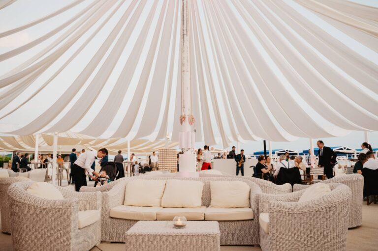 Cream rattan sofas under a white draped marquee on the beach, guests in black tie behind, Il Giorno Perfetto di Glenda Marradi