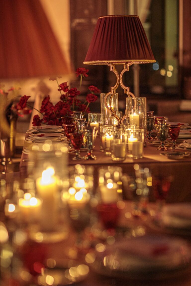 A red pleated lampshade over a candlelit table with red flowers and coloured glass, Il Giorno Perfetto di Glenda Marradi