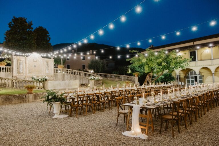 Long wooden tables laid in a villa courtyard under festoon lights at blue hour, Il Giorno Perfetto di Glenda Marradi