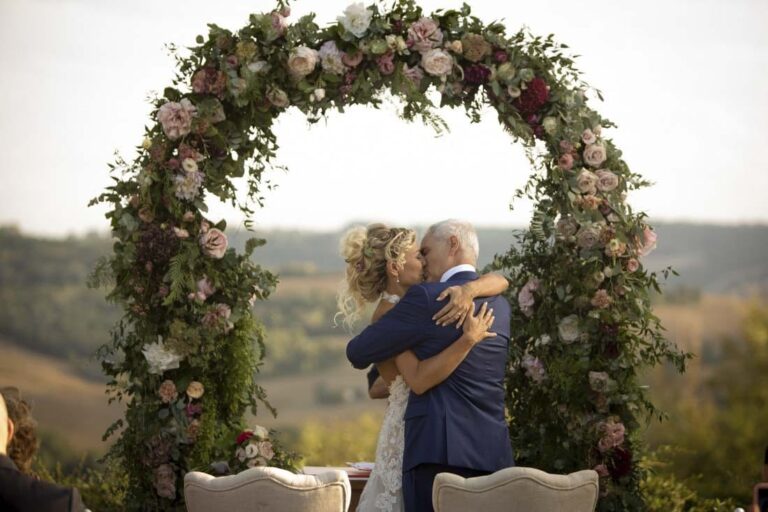 A couple embrace under a garden-rose arch at the end of their civil ceremony, rolling hills behind, Il Mio Matrimonio